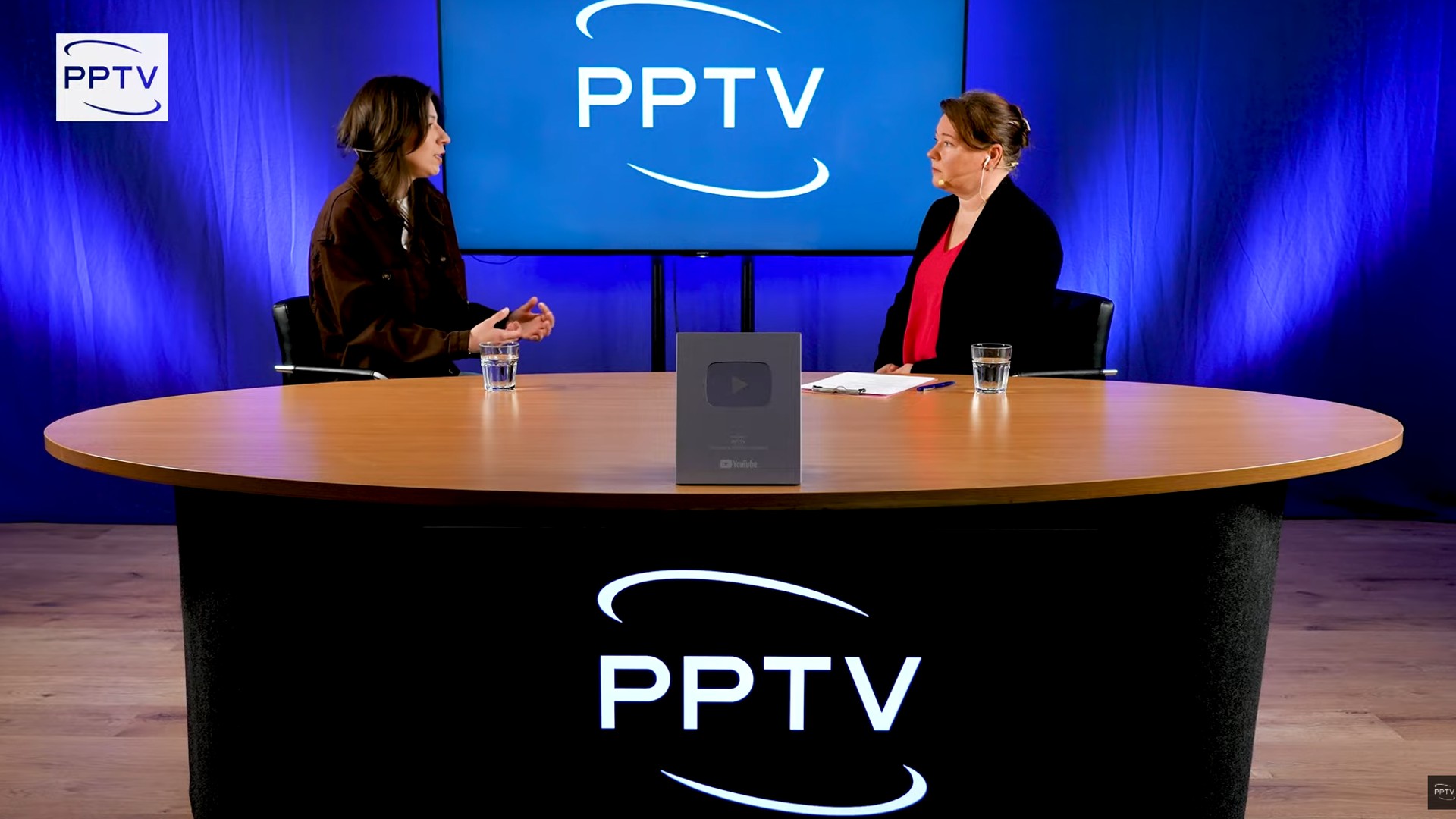 Two women sit at a round studio table with a large PPTV logo on a blue backdrop and a YouTube plaque on the table centerpiece, appearing to record or interview.