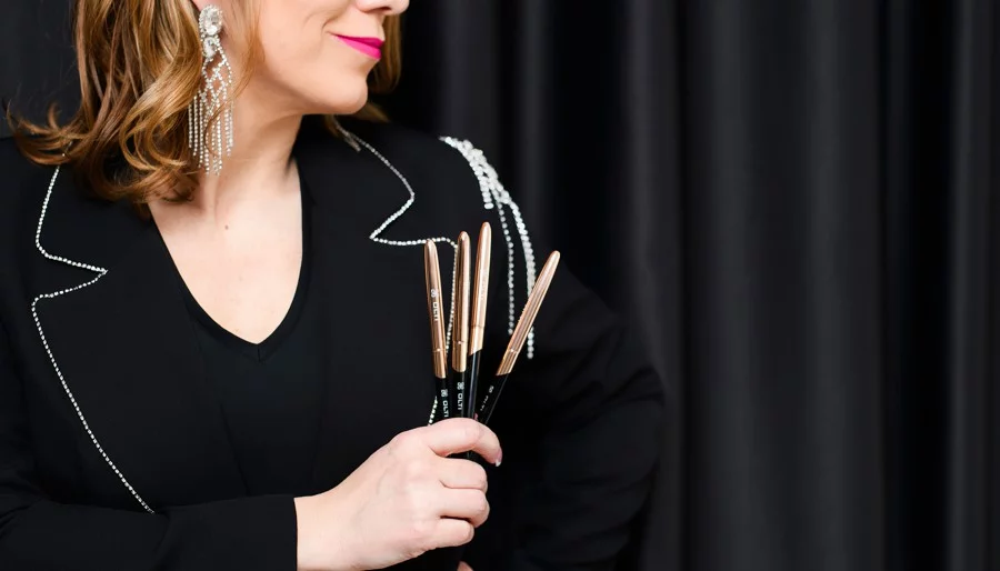 Woman in a black blazer with rhinestone trim holds several makeup brushes against a dark backdrop