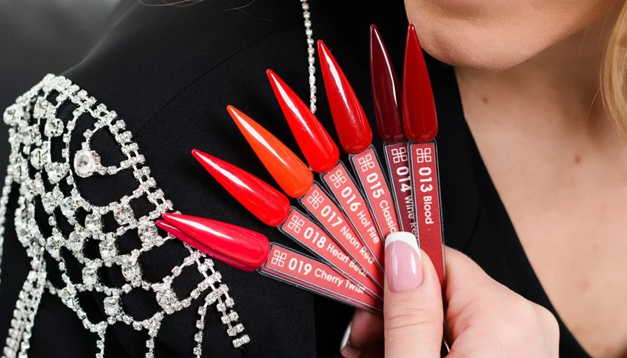 Close-up of glossy red lipstick shade swatches fanned with codes (e.g., 013 Blood, 019 Cherry Twist) held near a hand on a black embellished garment.