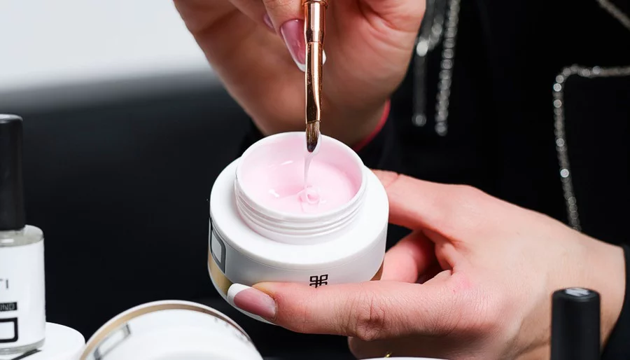 Close-up of a nail technician’s hands dipping a brush into a pink gel in a white jar for a manicure