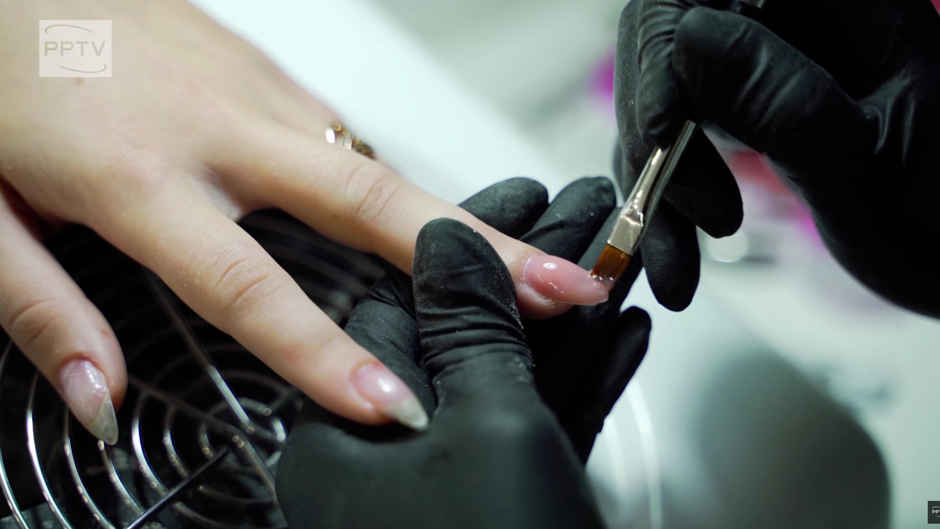 Close-up of a nail technician applying pink gel polish to a client's fingernail with a brush, wearing black gloves.
